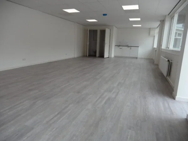 Lege, lichte ruimte met grijze laminaatvloer, systeemplafond en een kitchenette aan de achterzijde, gelegen aan de Jacques Perkstraat.